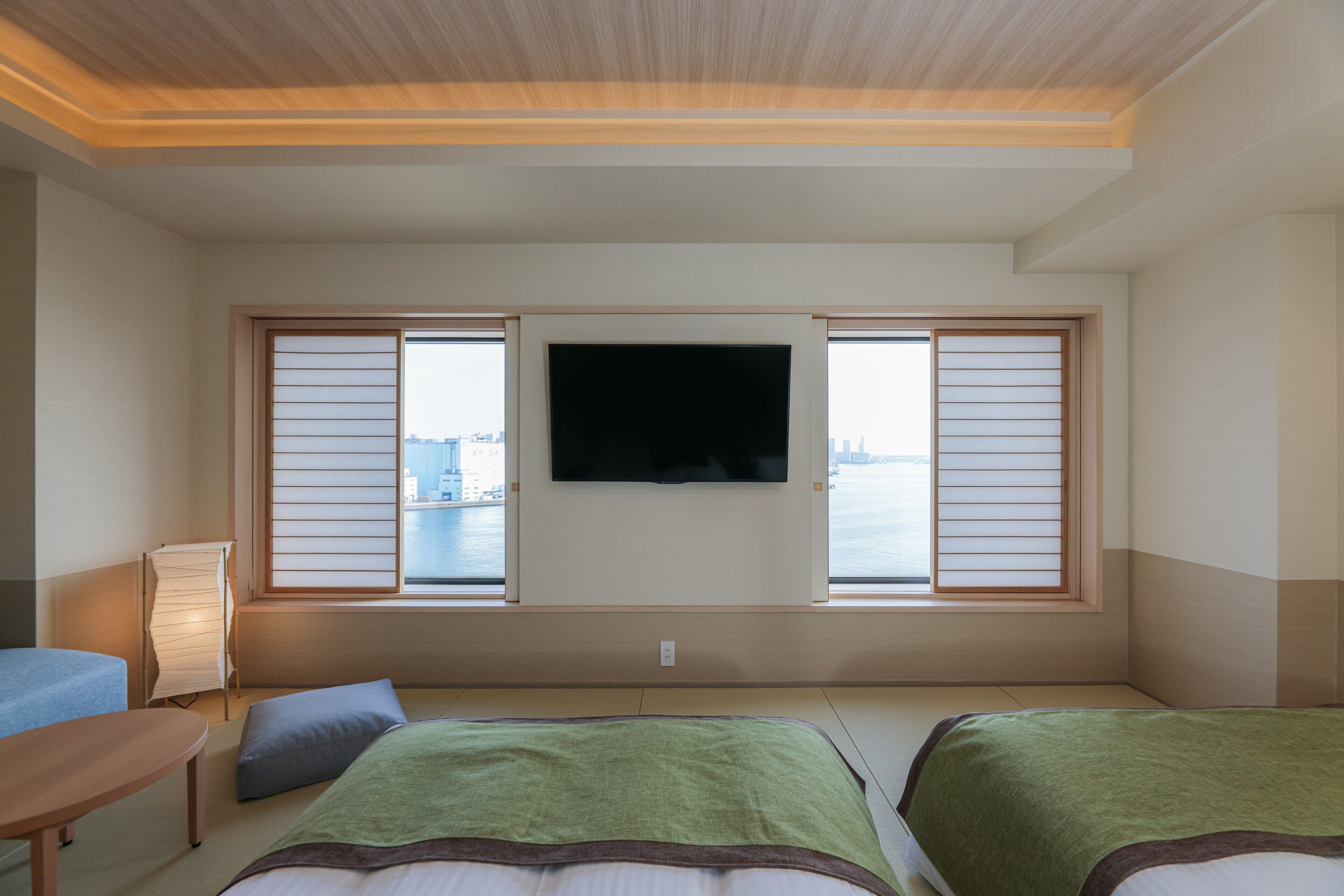 A hotel room with a large window, a flat-screen TV, two beds, a bedside table, and a lamp.