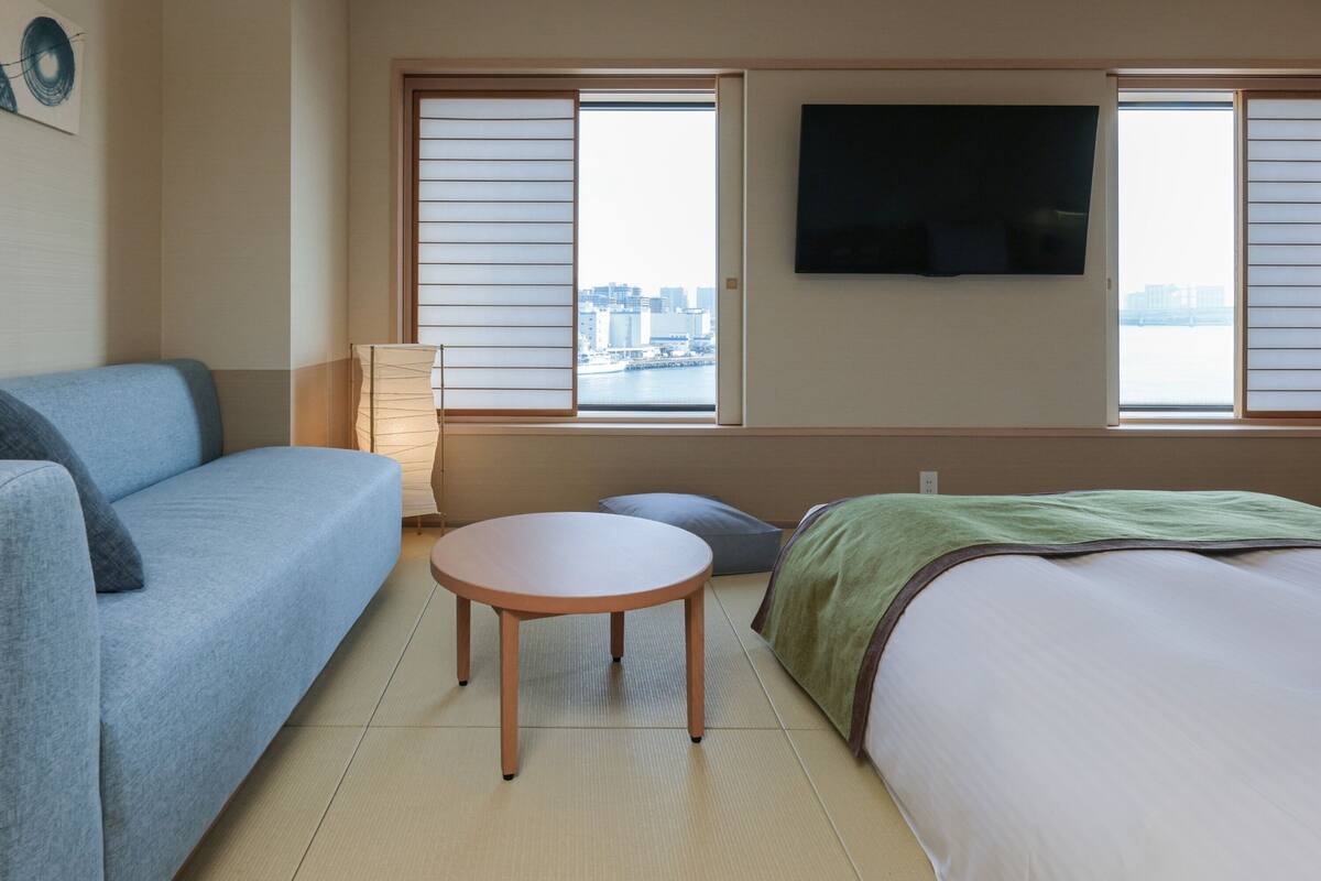 Une chambre d’hôtel avec un lit, un canapé, une petite table ronde, une télévision et une vue sur le paysage urbain par les fenêtres.