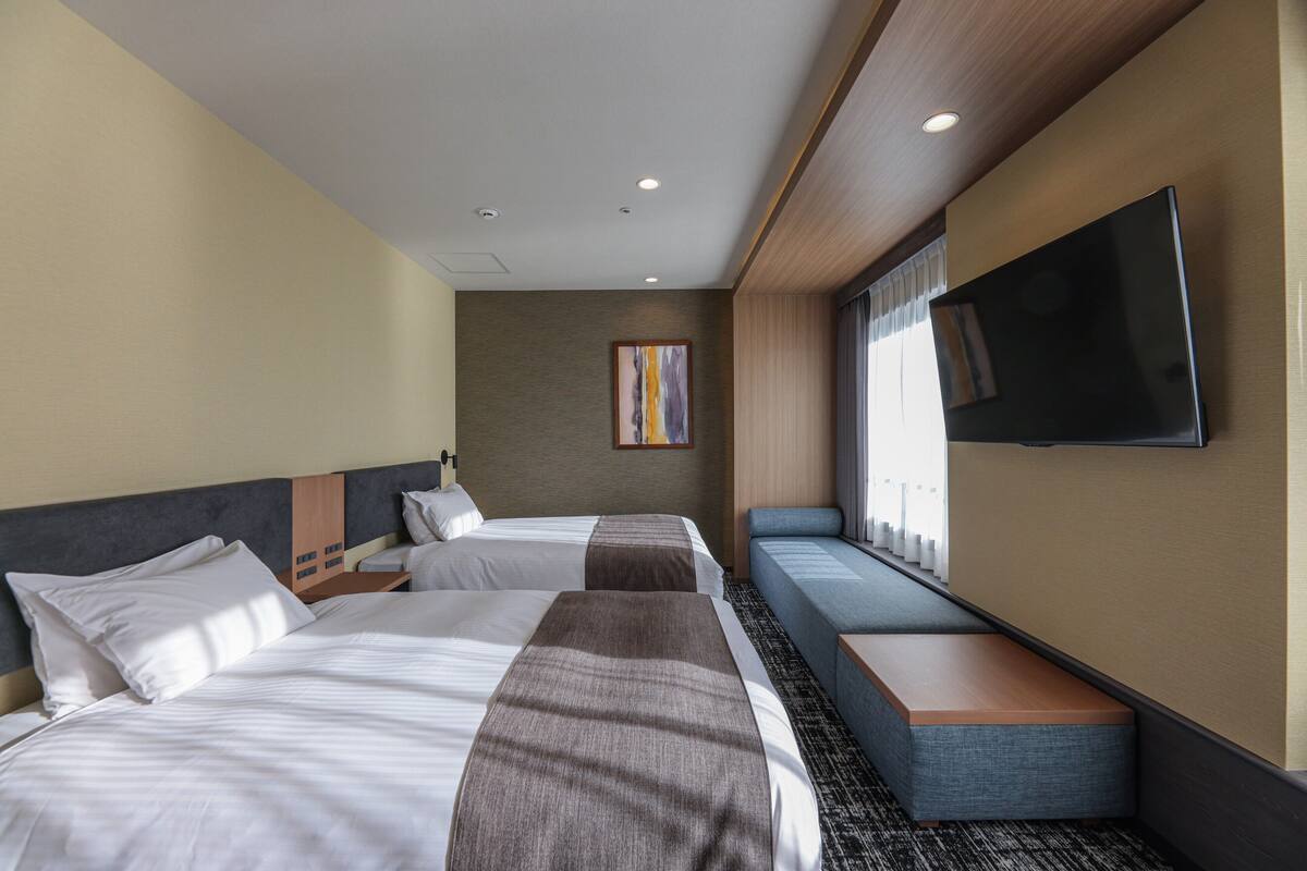 Une chambre d’hôtel avec deux lits, un téléviseur fixé au mur et un coin salon près de la fenêtre.