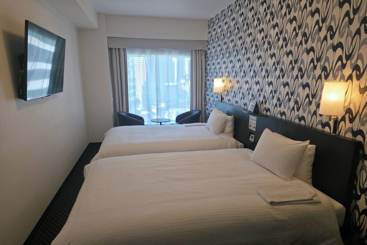 Une chambre d’hôtel avec deux lits, une télévision, une fenêtre avec des rideaux, un mur décoré et une petite table avec des chaises.