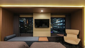 Ein Hotelzimmer mit zwei Betten, einem Fernseher, einem Sessel und Blick auf eine nächtliche Stadtlandschaft.