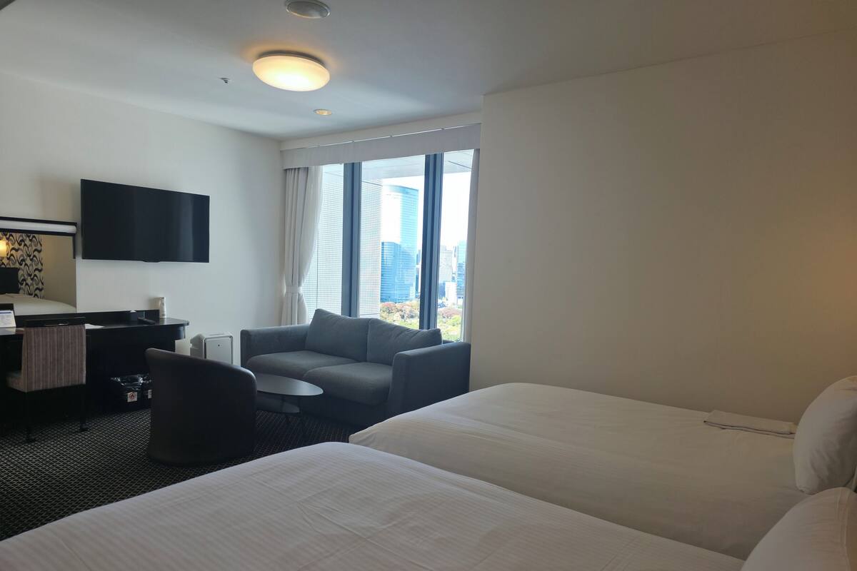 Une chambre d’hôtel avec deux lits, un bureau avec une chaise, un canapé, un téléviseur à écran plat fixé au mur et de grandes fenêtres donnant sur la ville.