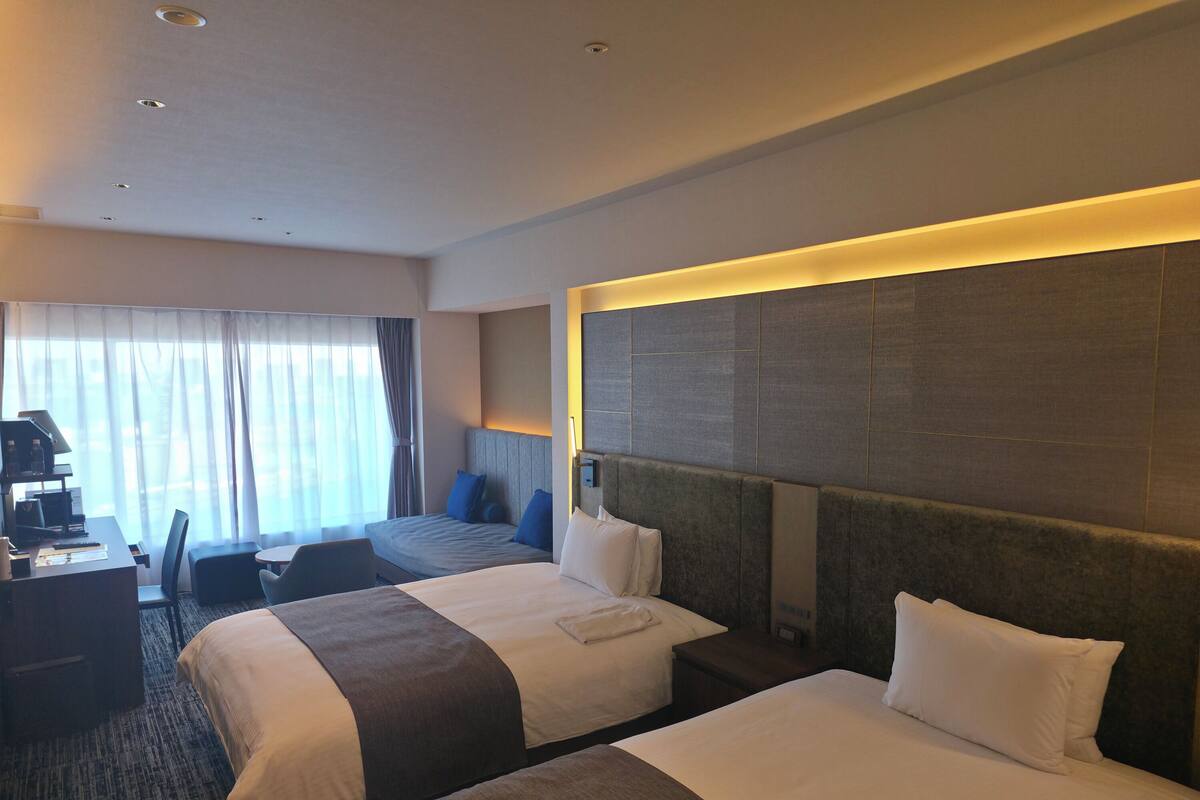 Une chambre d’hôtel avec deux lits, un bureau avec une lampe, une chaise et une grande fenêtre avec des rideaux.