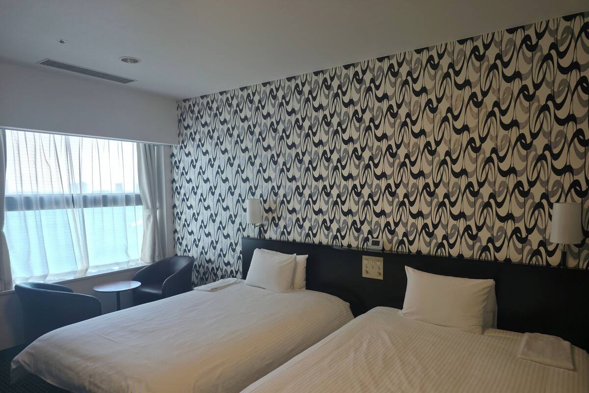 Une chambre d’hôtel avec deux lits, un bureau, une chaise et une fenêtre avec des rideaux.