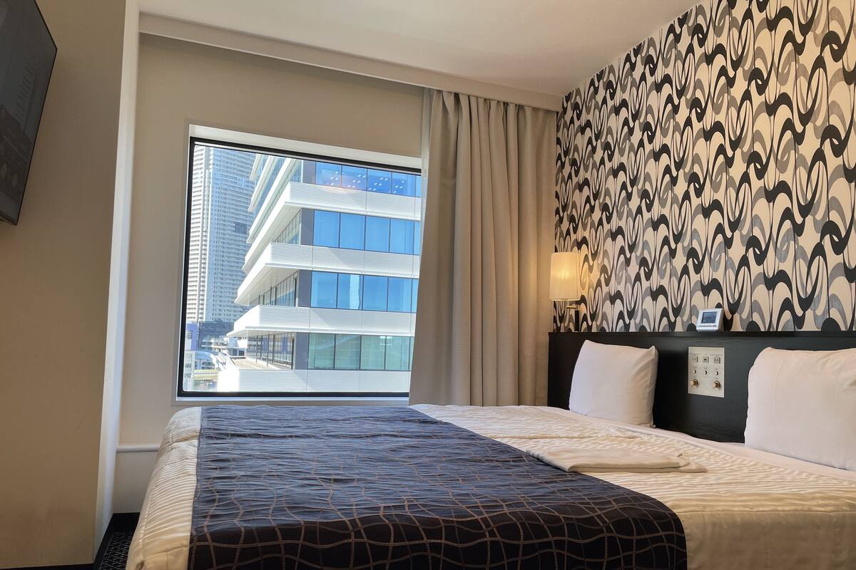 Une chambre d’hôtel avec un grand lit, une fenêtre donnant sur un immeuble moderne, un mur décoré d’un motif et une table de chevet avec une lampe.