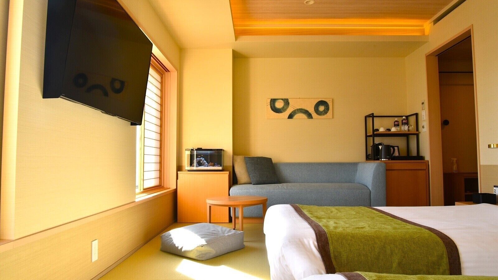 Quarto de hotel com cama, sofá, mesa pequena, aquário e televisão fixada na parede.