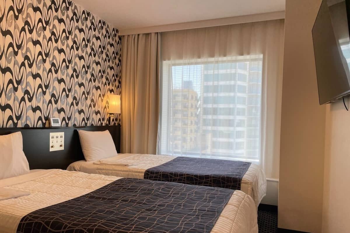 Une chambre d’hôtel avec deux lits, un mur décoré de motifs, une fenêtre donnant sur des immeubles et un téléviseur.