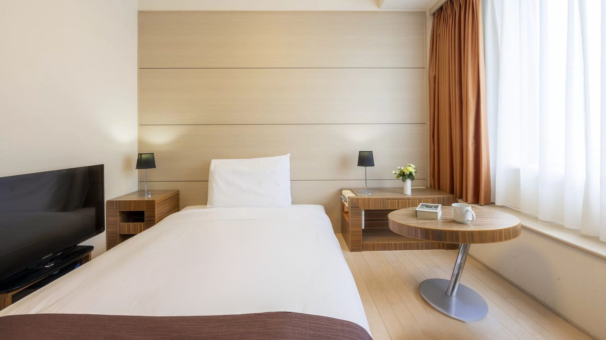A hotel room with a bed, a TV, a bedside table with a lamp, a small table with a plant, and a window with curtains.