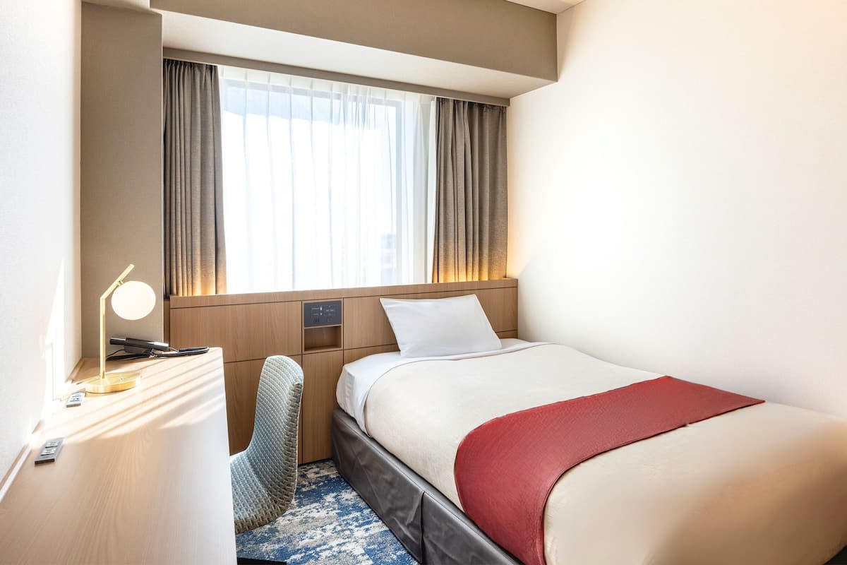 Une chambre d’hôtel avec un lit, un bureau avec une lampe, une chaise et une fenêtre avec des rideaux.