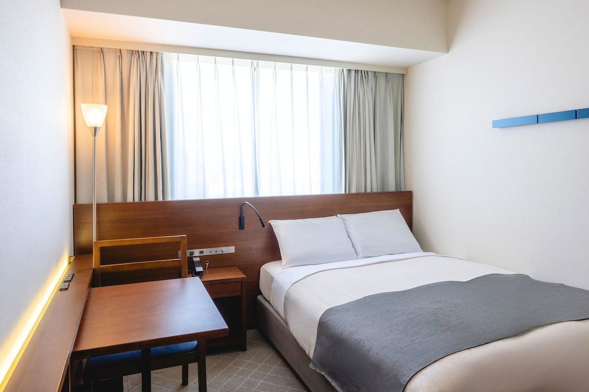 Une chambre d’hôtel avec un lit, un bureau, une chaise, une lampe et une fenêtre avec des rideaux.