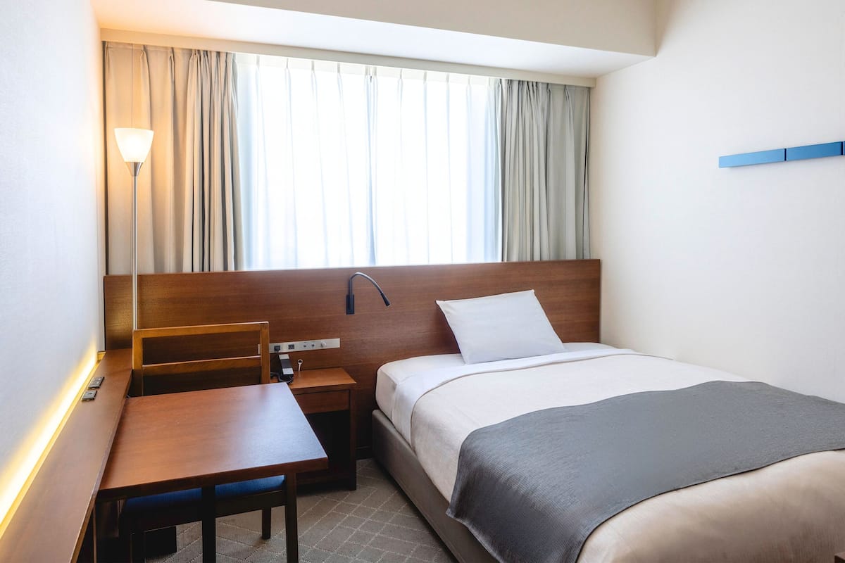 Une chambre d’hôtel avec une tête de lit en bois, un lit recouvert d’une couverture grise, un bureau avec une chaise, une table de chevet et une fenêtre avec des rideaux.
