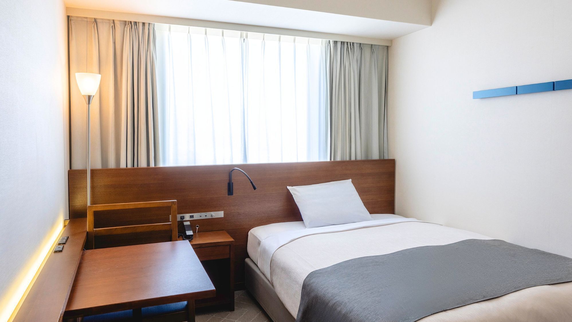 A hotel room with a bed, bedside table, lamp, and a window with curtains.