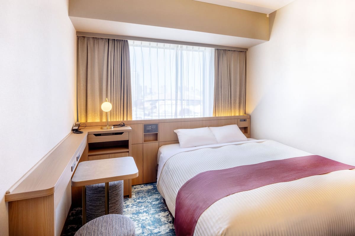 Une chambre d’hôtel avec un lit, un bureau, une chaise, une fenêtre avec des rideaux et une lampe.
