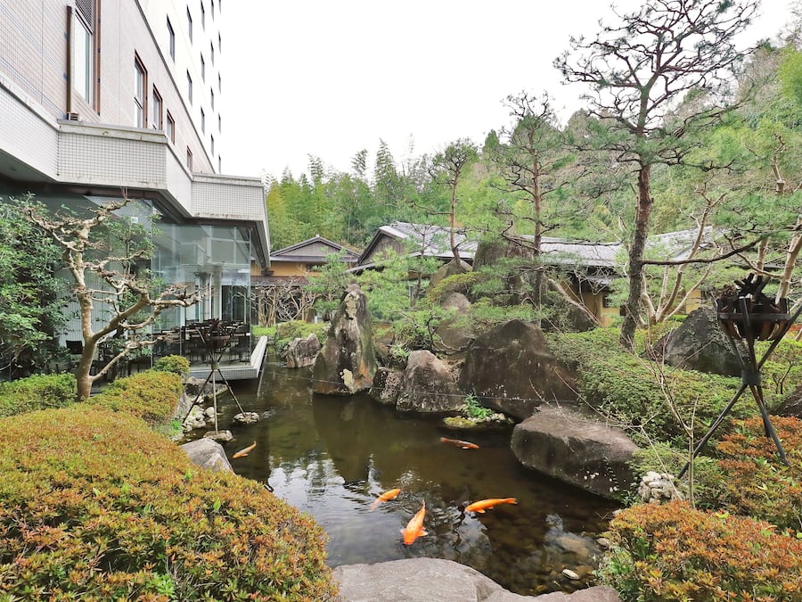 Un estanque de carpas koi rodeado de rocas y follaje, con un edificio al fondo.