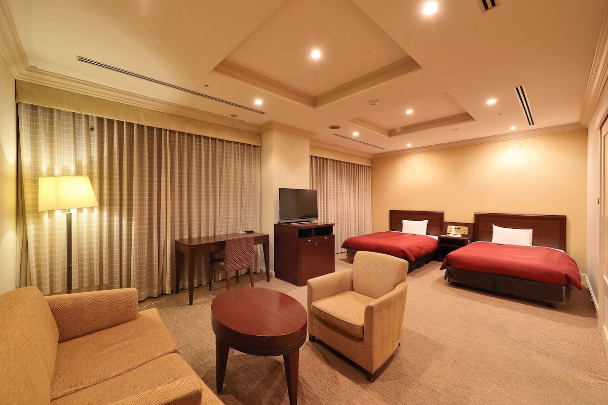A hotel room with two beds, a desk, a chair, a TV, and a lamp.