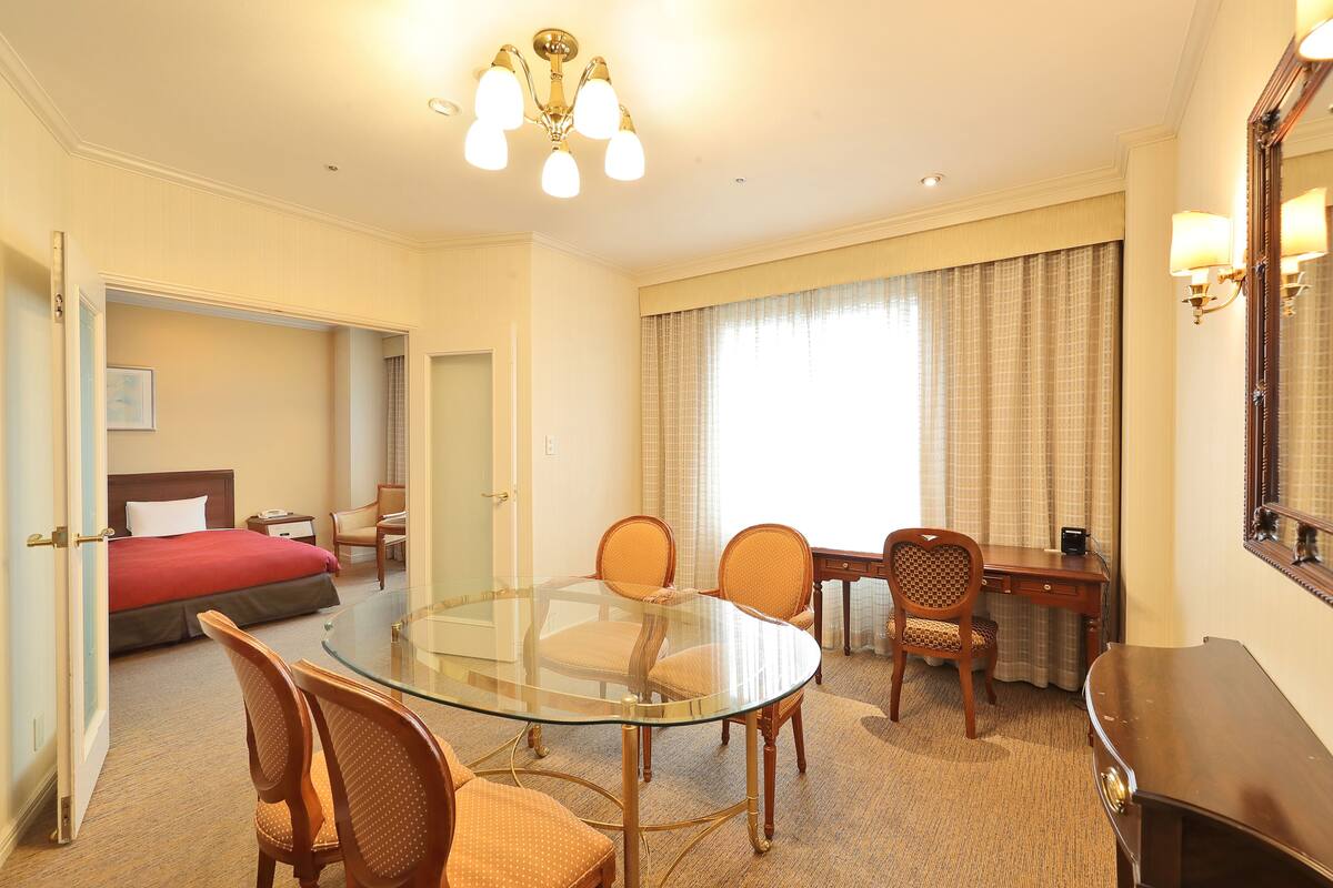 A hotel room with a glass table, four chairs, a desk, a bed, a mirror, and a chandelier.