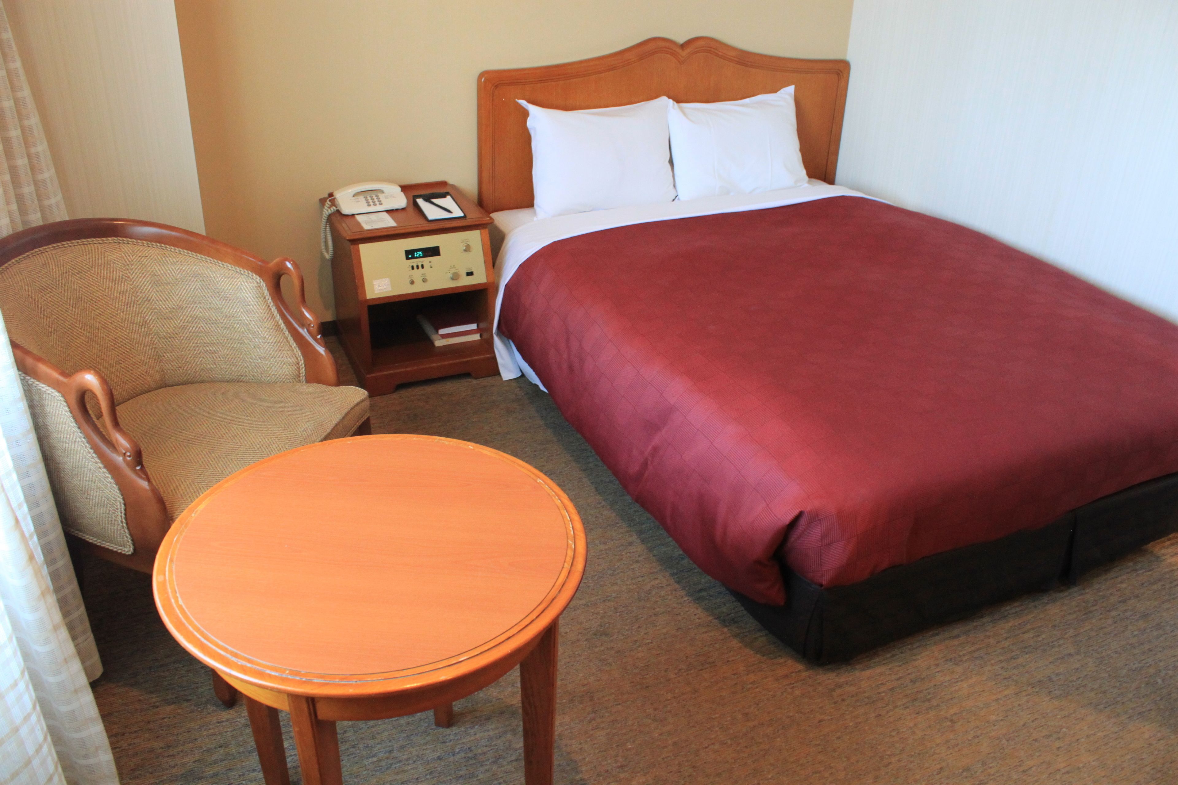 Ett hotellrum med en säng, ett träbord vid sängen med en telefon, en stol och ett litet bord.