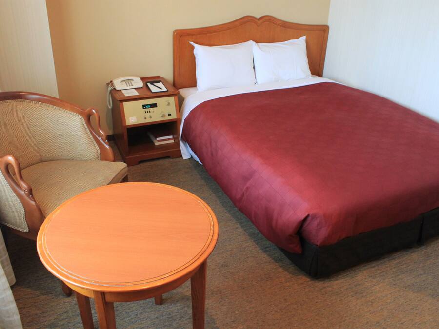 A hotel room with a bed, a wooden nightstand with a telephone, a chair, and a small table.