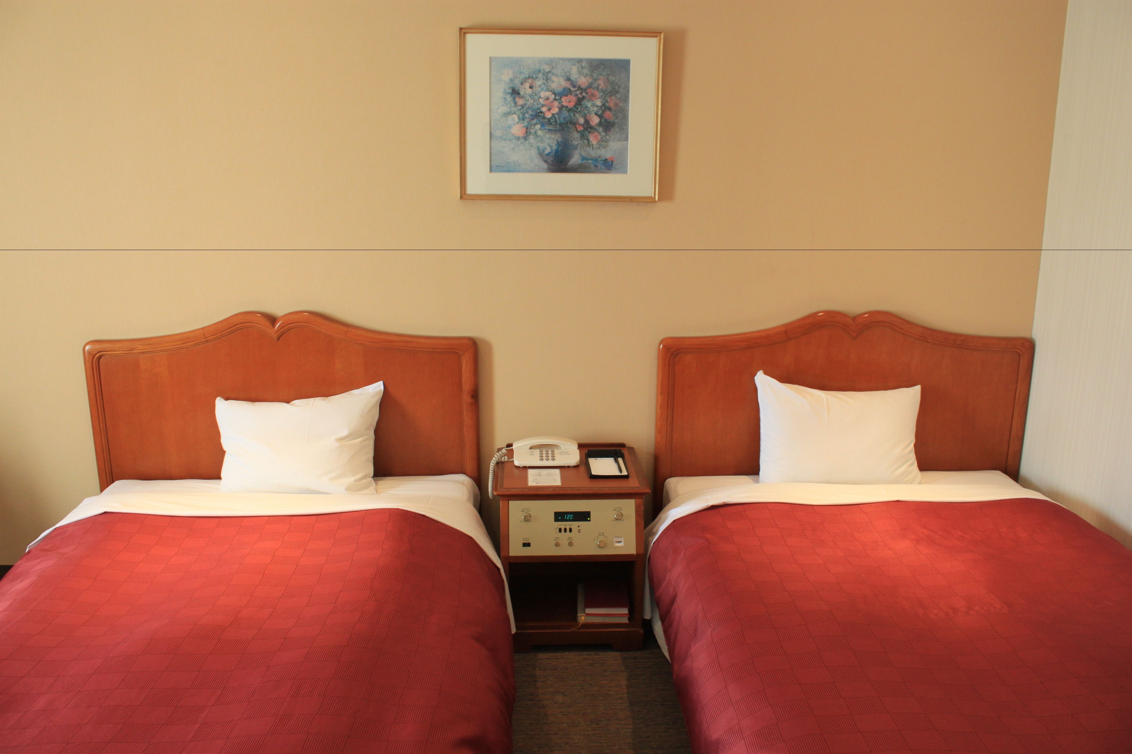 Une chambre d’hôtel avec deux lits, chacun doté d’une tête de lit en bois, d’oreillers blancs et d’une literie rouge. Une table de chevet se trouve entre les lits, avec un téléphone et une petite lampe. Un tableau encadré est accroché au mur au-dessus des lits.