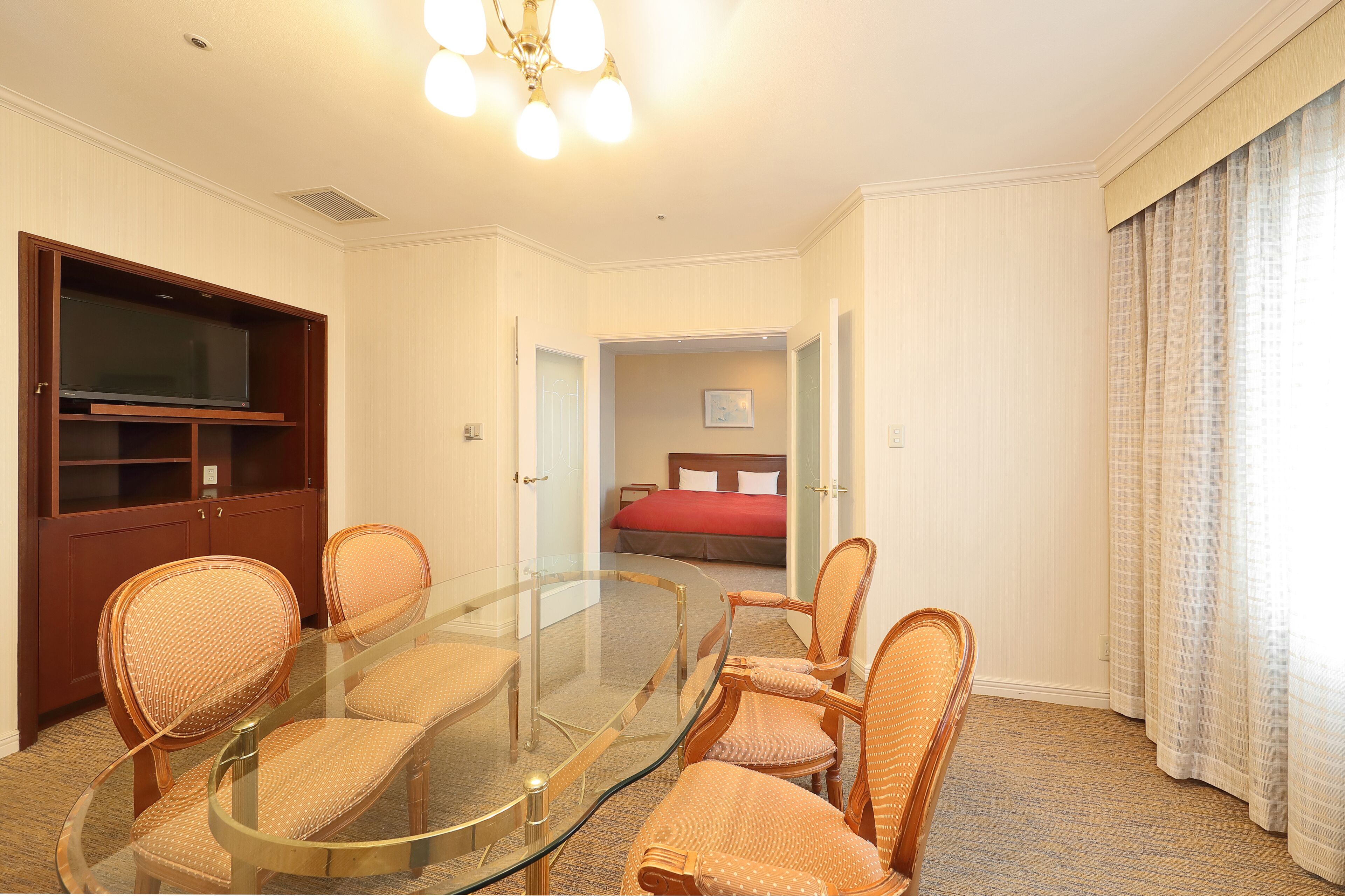 Una camera d'hotel con un tavolo da pranzo in vetro, sedie in vimini, un armadio in legno e un letto con biancheria rossa.