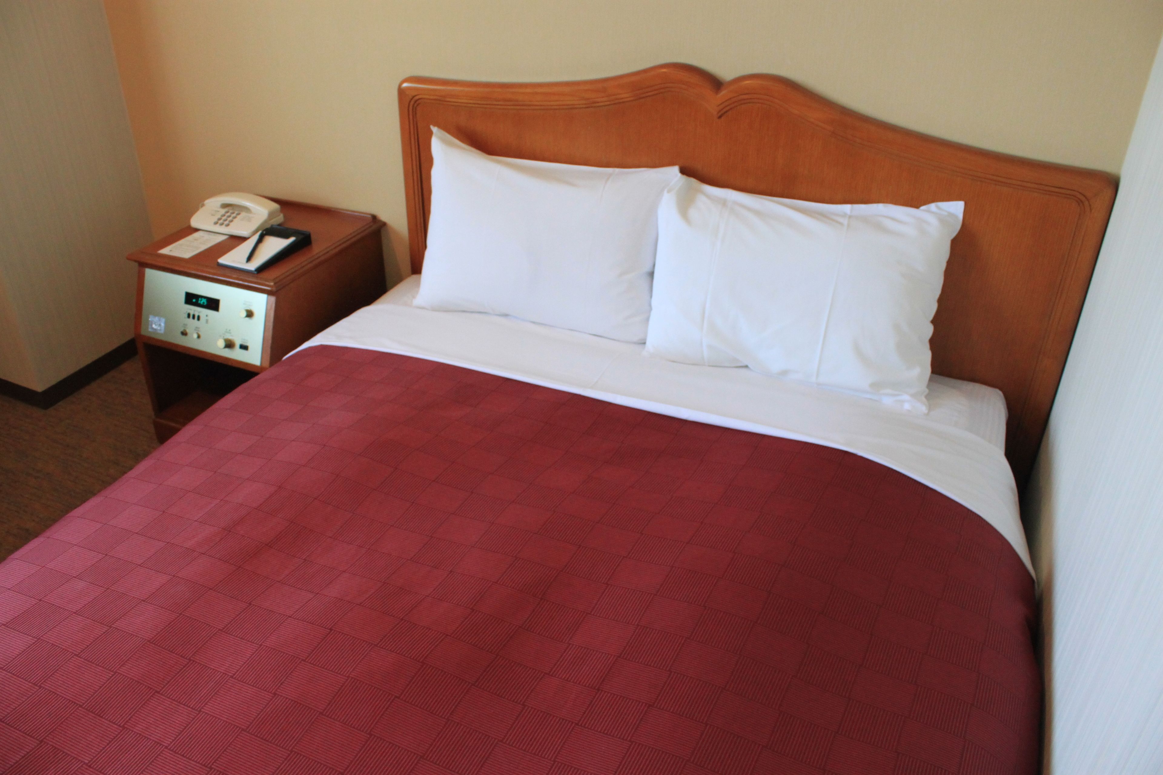 A hotel room with a wooden headboard, a red quilted bedspread, a white pillow, and a bedside table with a phone and a notepad.
