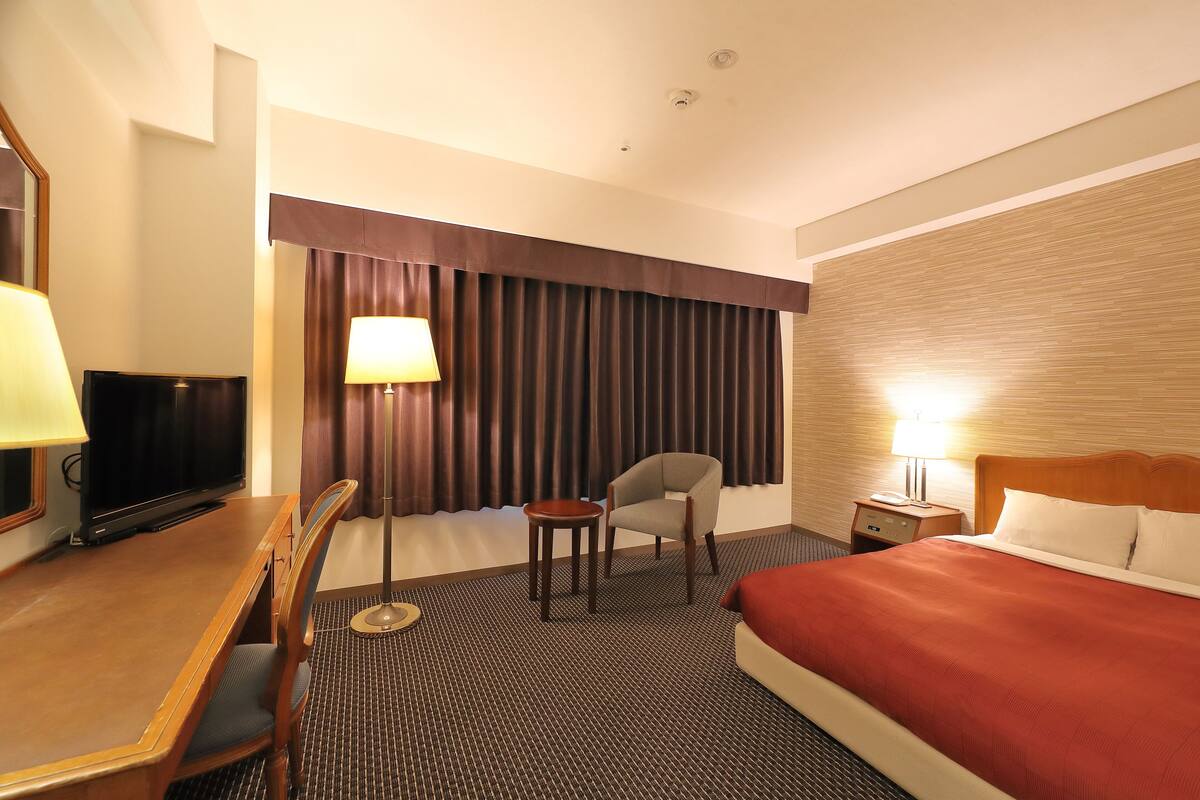 A hotel room with a bed, a desk with a chair, a television, a lamp, and a nightstand with a lamp.
