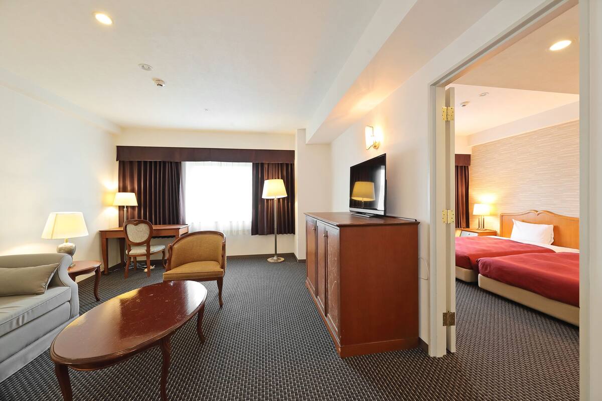 A hotel room with a sofa, a small table, a desk with a lamp, a television on a cabinet, and a bed with red bedding.