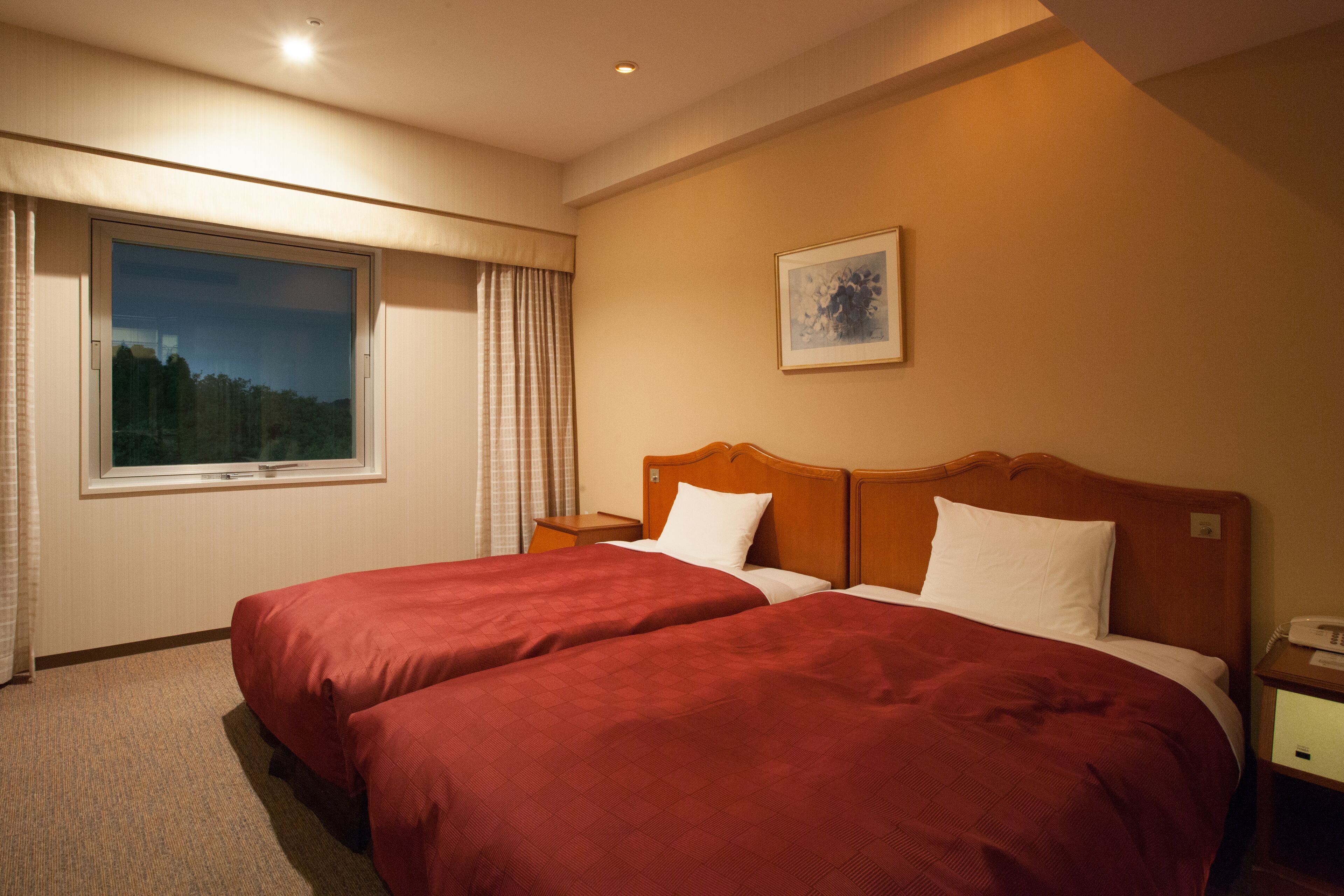 A hotel room with two beds, a window, a bedside table, and a wall-mounted painting.