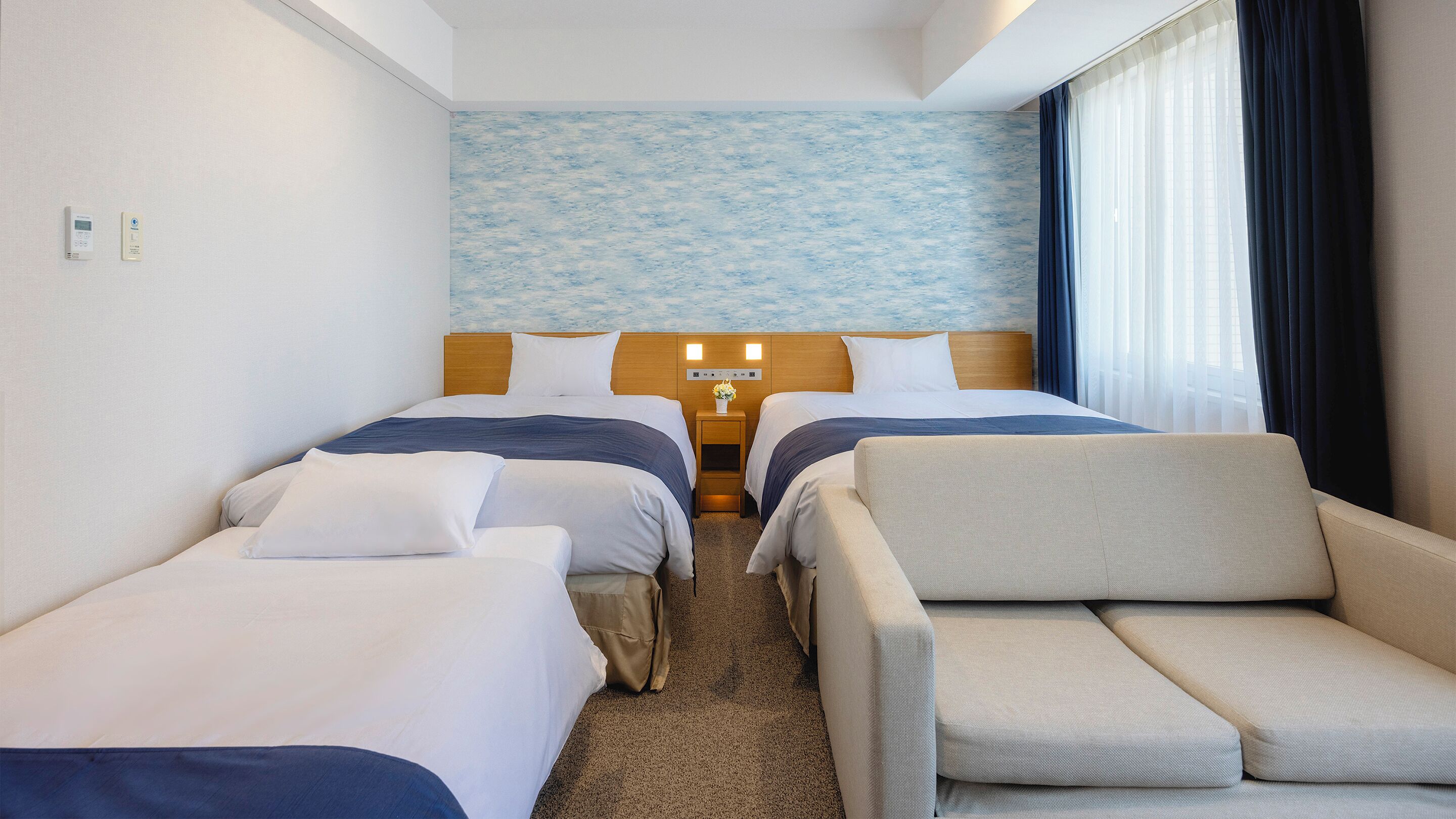 Une chambre d’hôtel avec deux lits, un canapé, une table de chevet avec une lampe et un panneau de commande fixé au mur.