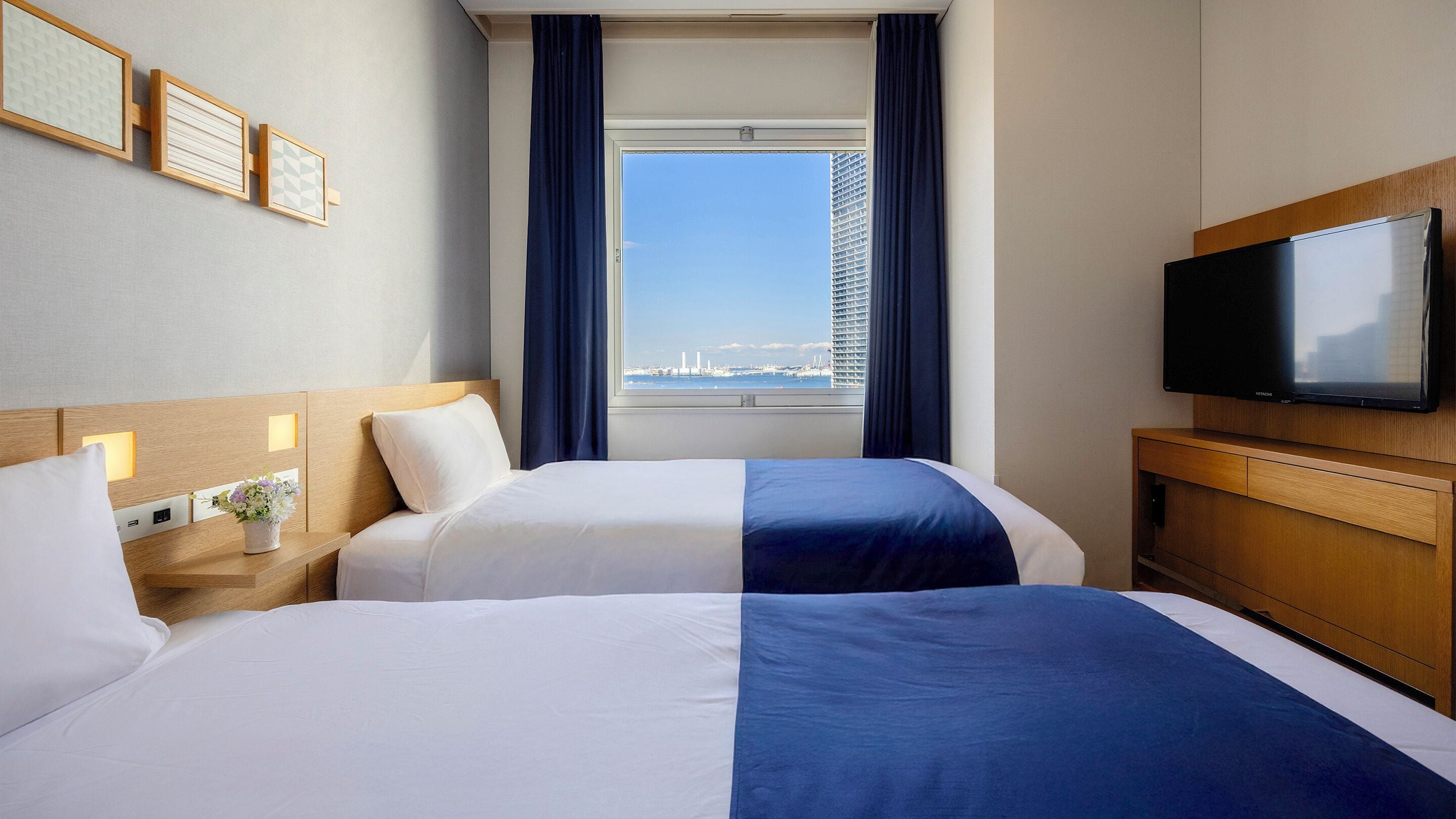 Ein Hotelzimmer mit zwei Betten, einem Fernseher, einem Fenster mit Blick auf die Stadt und einer Wand mit gerahmten Bildern.