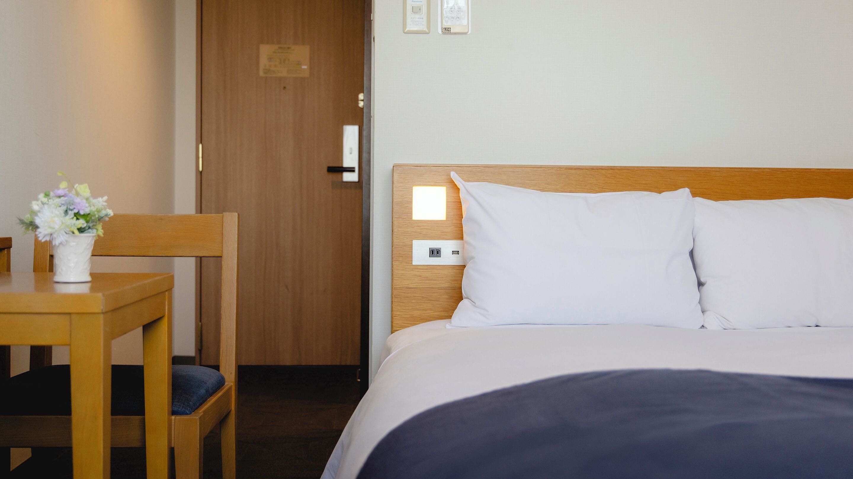 A hotel room with a wooden headboard, a bed with white linens, a small table with a vase of flowers, and a wooden chair.