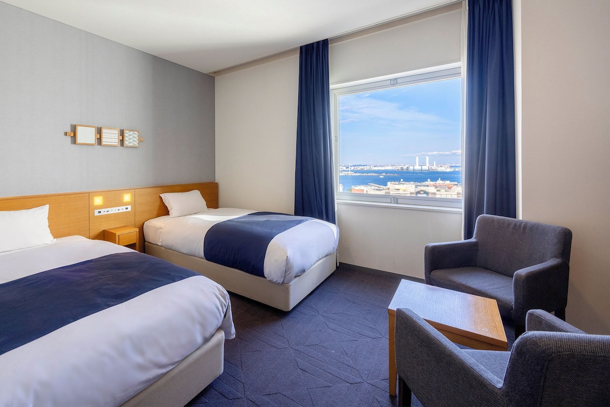 A hotel room with two beds, a desk, a chair, and a view of a cityscape.