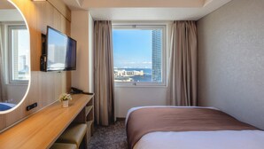 A hotel room with a bed, a desk with a TV, a phone, a small vase with flowers, and a window with a city view. - Yokohama Sakuragicho Washington Hotel (Yokohama)