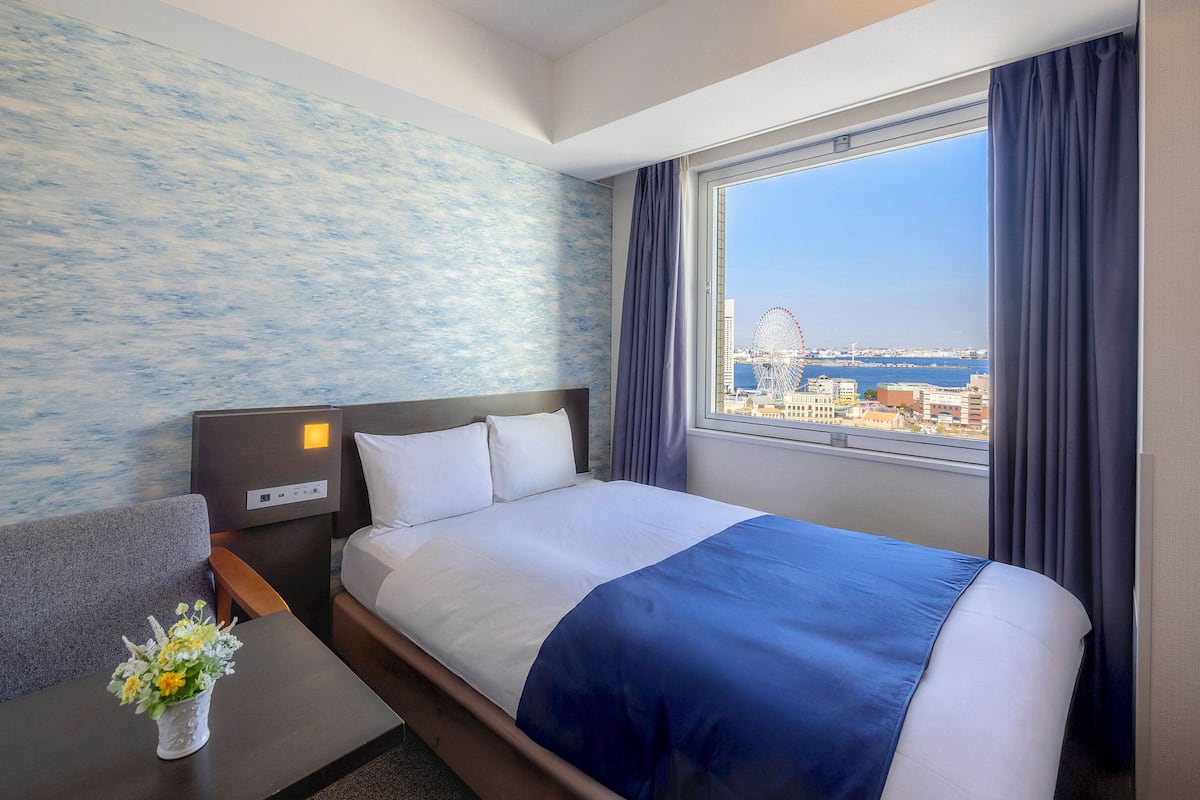 A hotel room with a bed, a desk, a chair, a nightstand, a window with a city view, and a wall with a textured pattern.