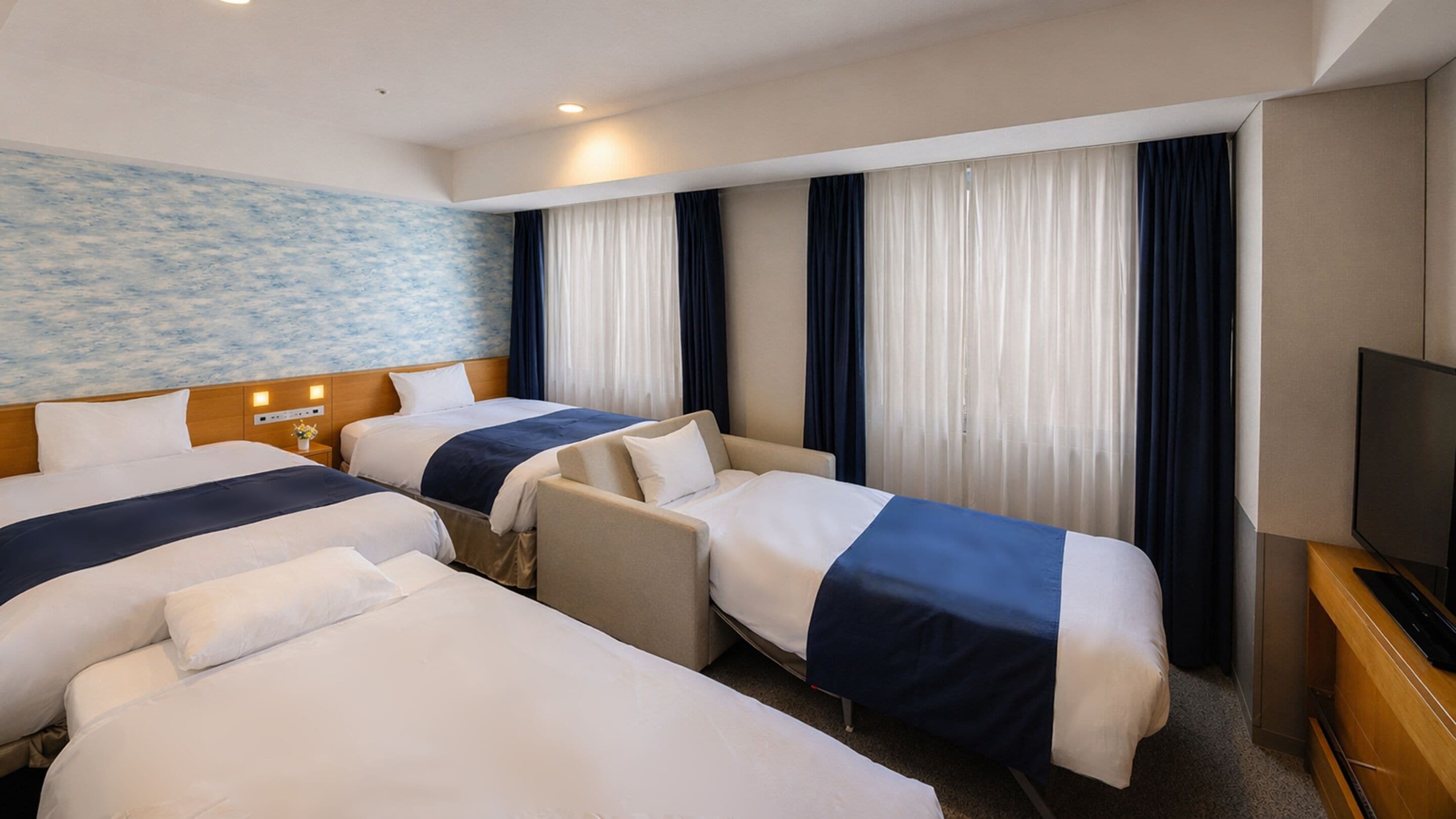 A hotel room with two beds, a sofa, a TV, and a wall with a blue pattern.