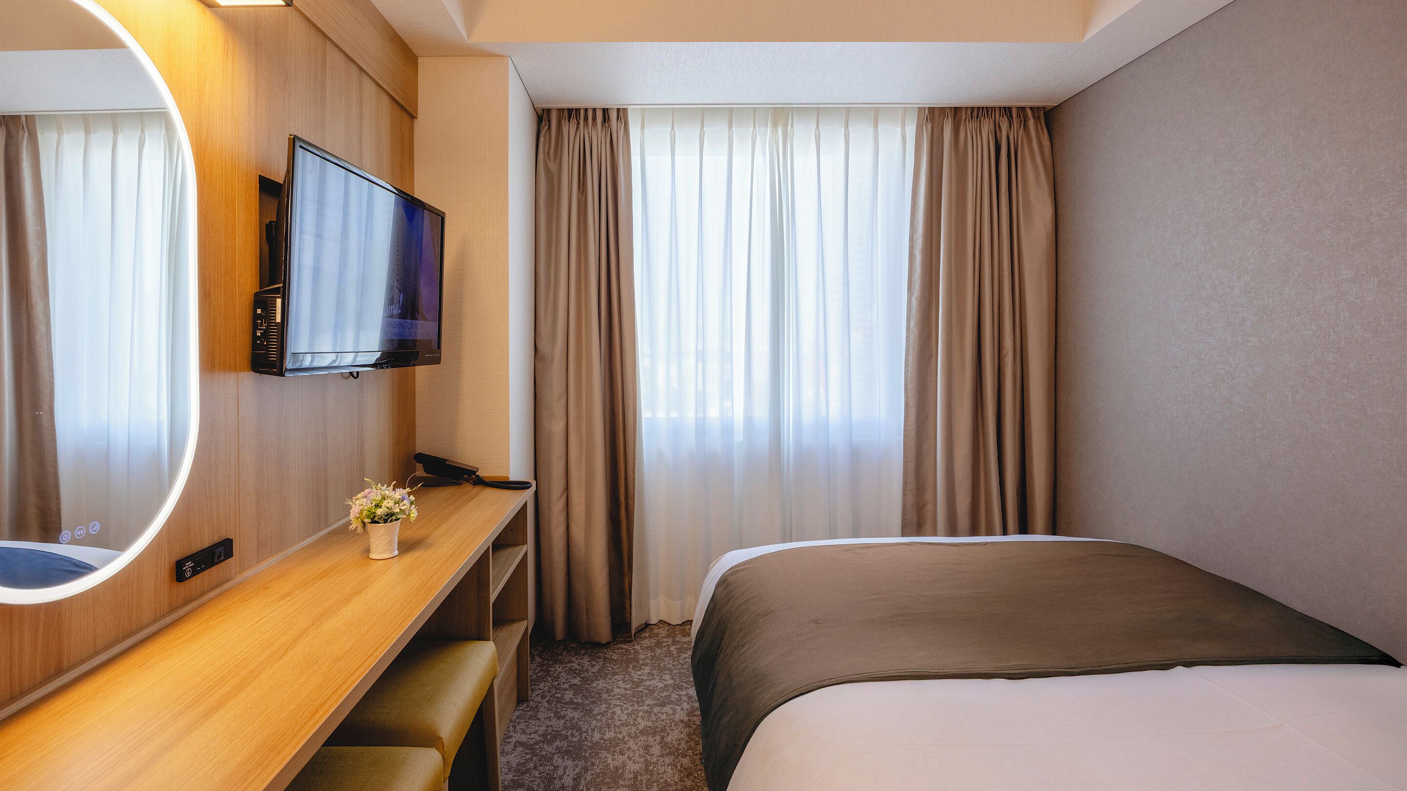 Ein Hotelzimmer mit Bett, Schreibtisch mit Fernseher, Spiegel und Vorhängen.