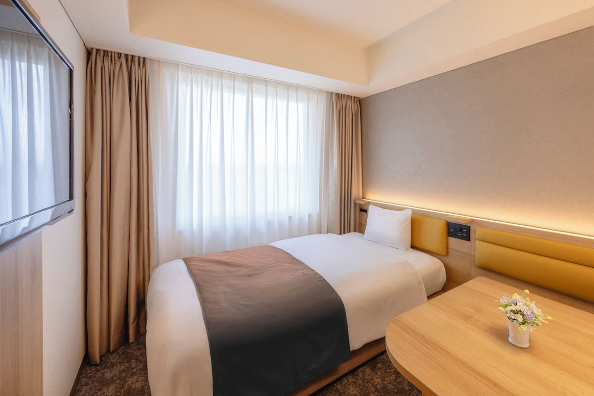 A hotel room with a bed, a bedside table with a vase of flowers, a flat-screen TV, and a window with curtains.