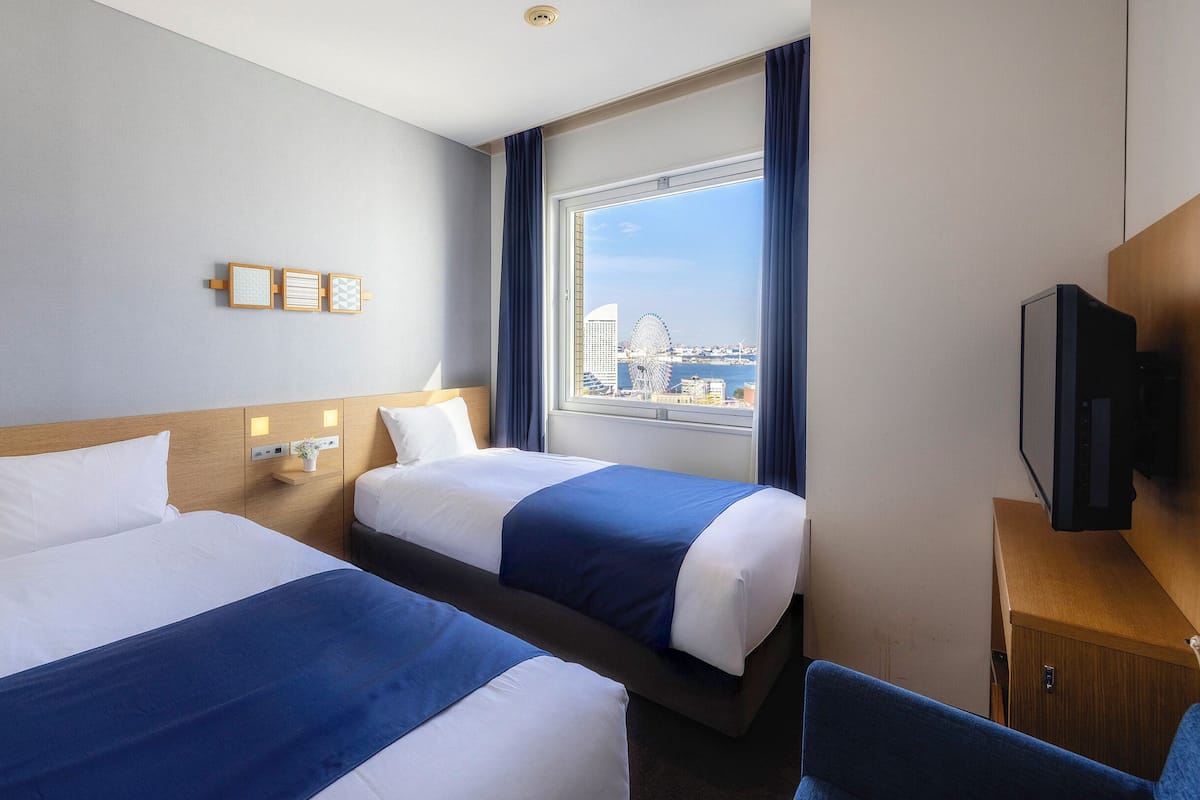 A hotel room with two beds, a TV, a desk, and a window with a city view.