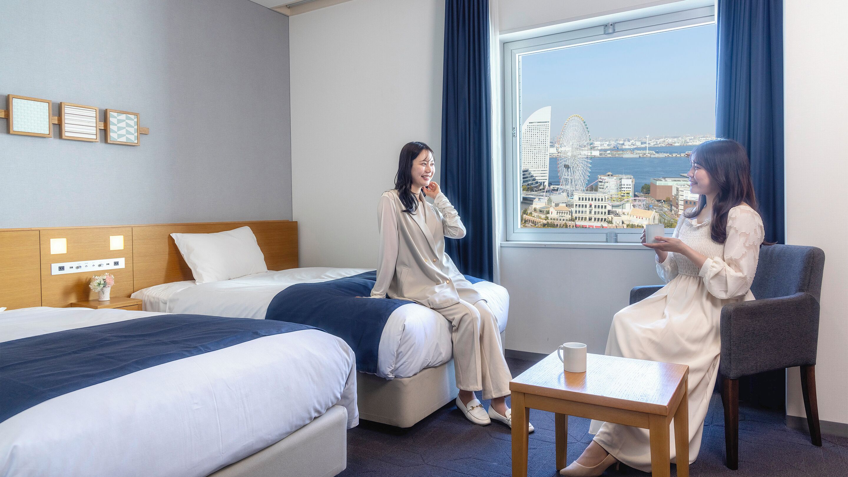 Habitación de hotel con dos camas, una mesita y vistas a un paisaje urbano.
