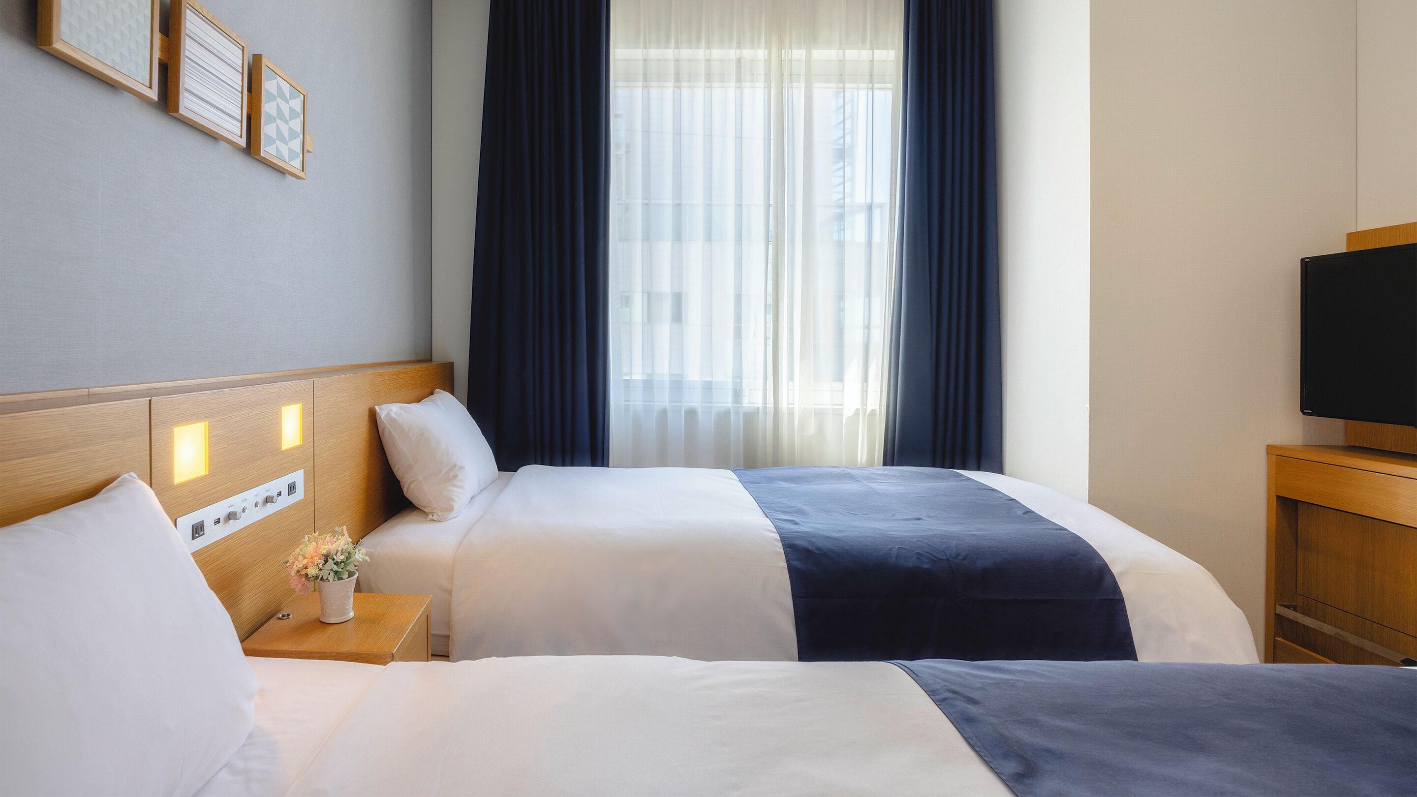 A hotel room with two beds, a TV, a nightstand with a vase of flowers, and a window with curtains.