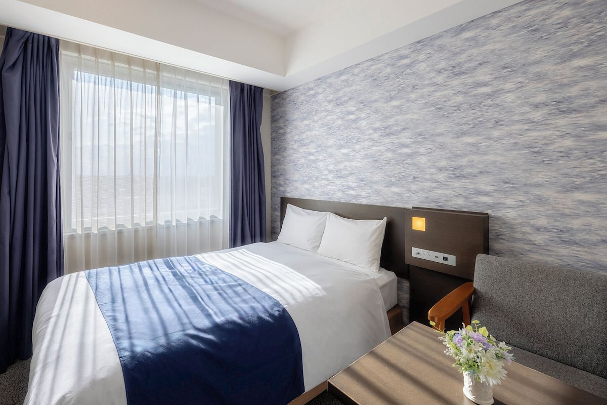 A hotel room with a bed, a desk, a chair, a nightstand, and a wall with a textured design.