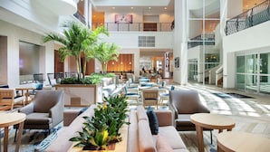 A spacious lobby with multiple seating areas, a central plant arrangement, and a staircase leading to an upper level.