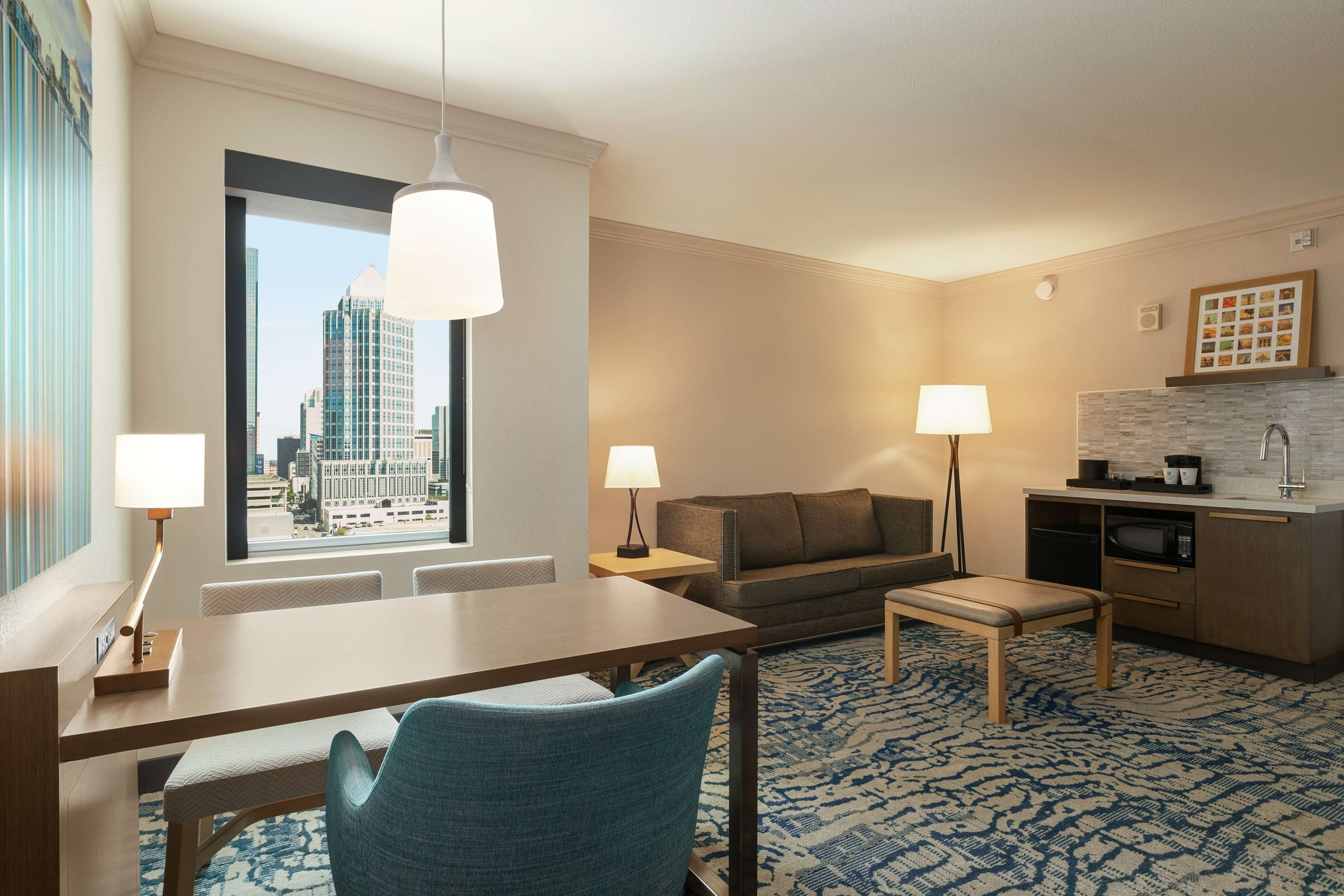 A hotel room with a dining table, two chairs, a sofa, a bench, a kitchen area with a microwave, and a large window offering a city view.