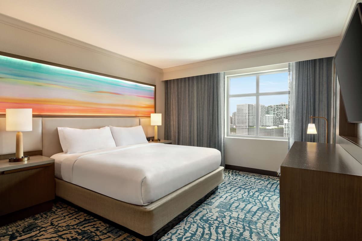 A hotel room with a large bed, two bedside tables with lamps, a large window with curtains, and a city view.