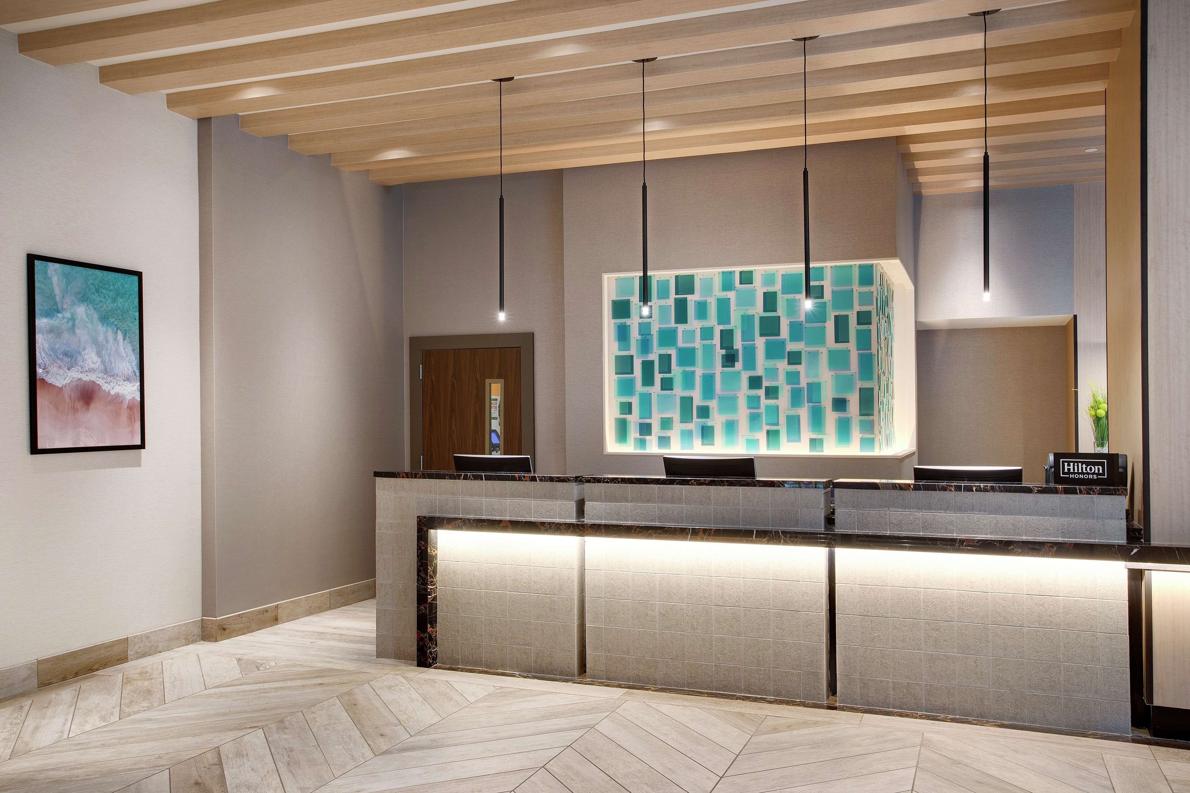 Uma área de recepção de hotel com design moderno, destacando-se um piso em padrão herringbone, um balcão de recepção com parede de mosaico de vidro e teto de madeira com luminárias pendentes.