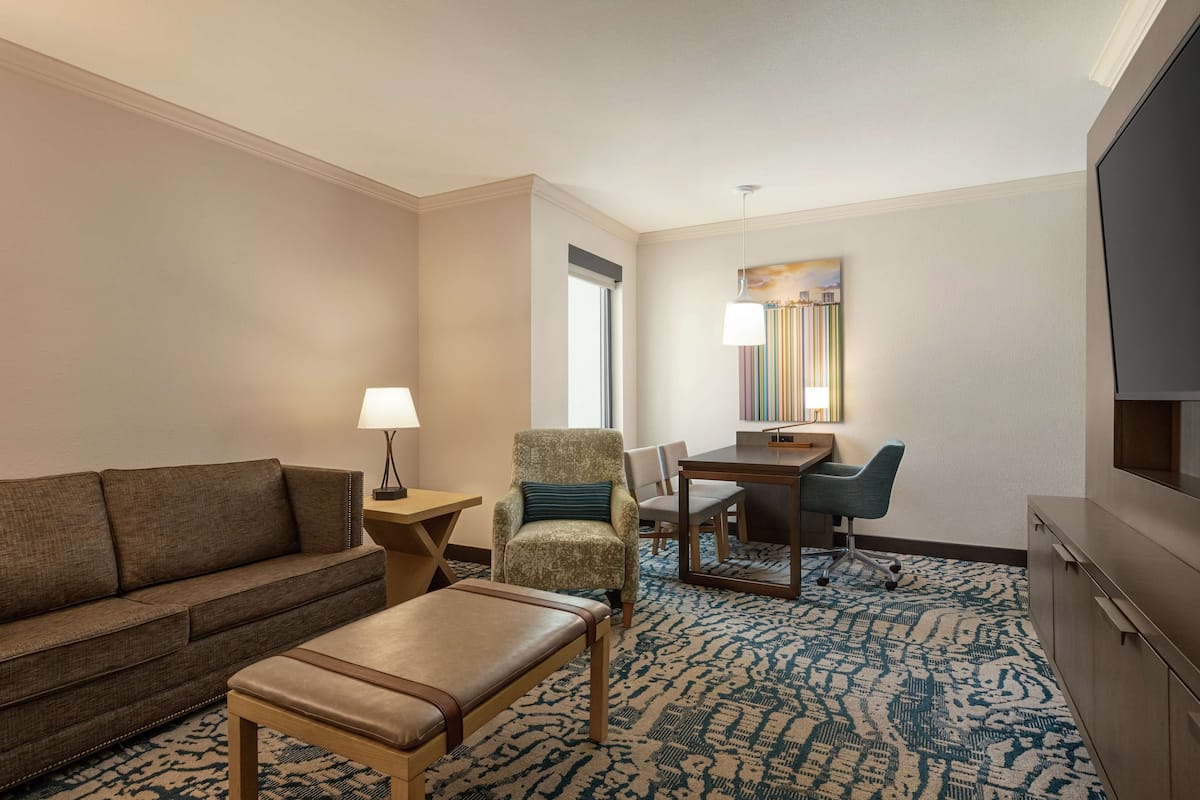 A hotel room with a sofa, armchair, ottoman, desk, chair, lamp, TV, and a patterned carpet.