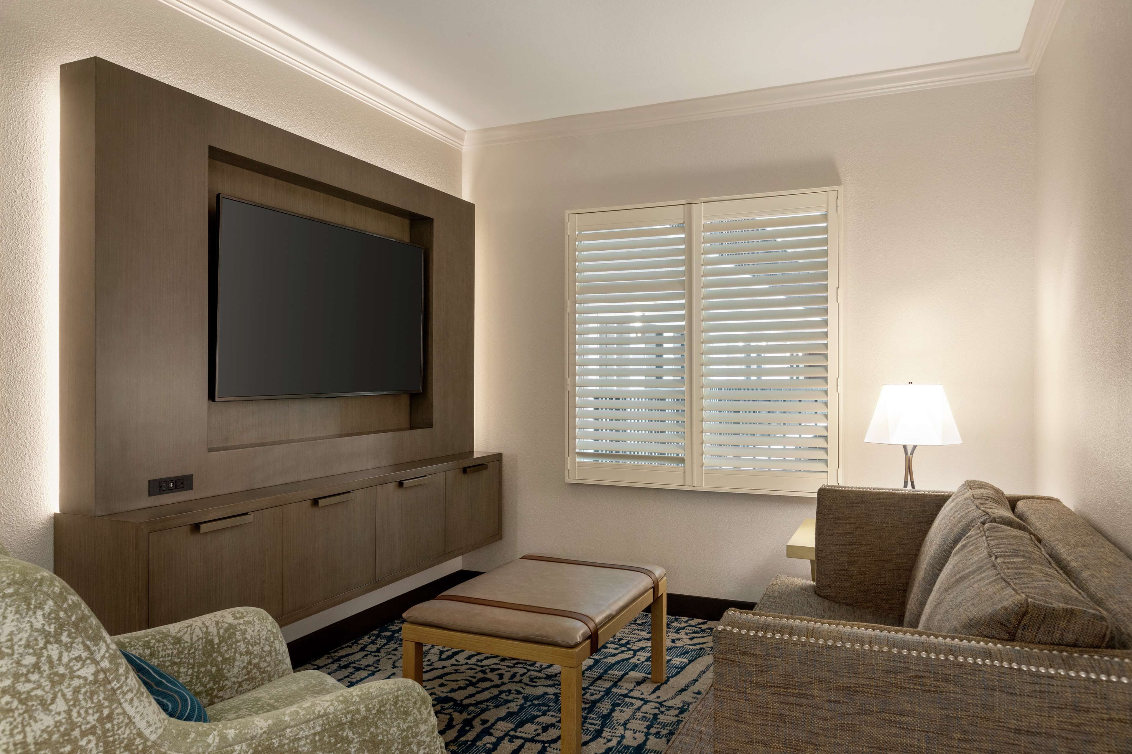 A living room with a flat-screen TV mounted on the wall, a sofa, a small table, and a window with blinds.