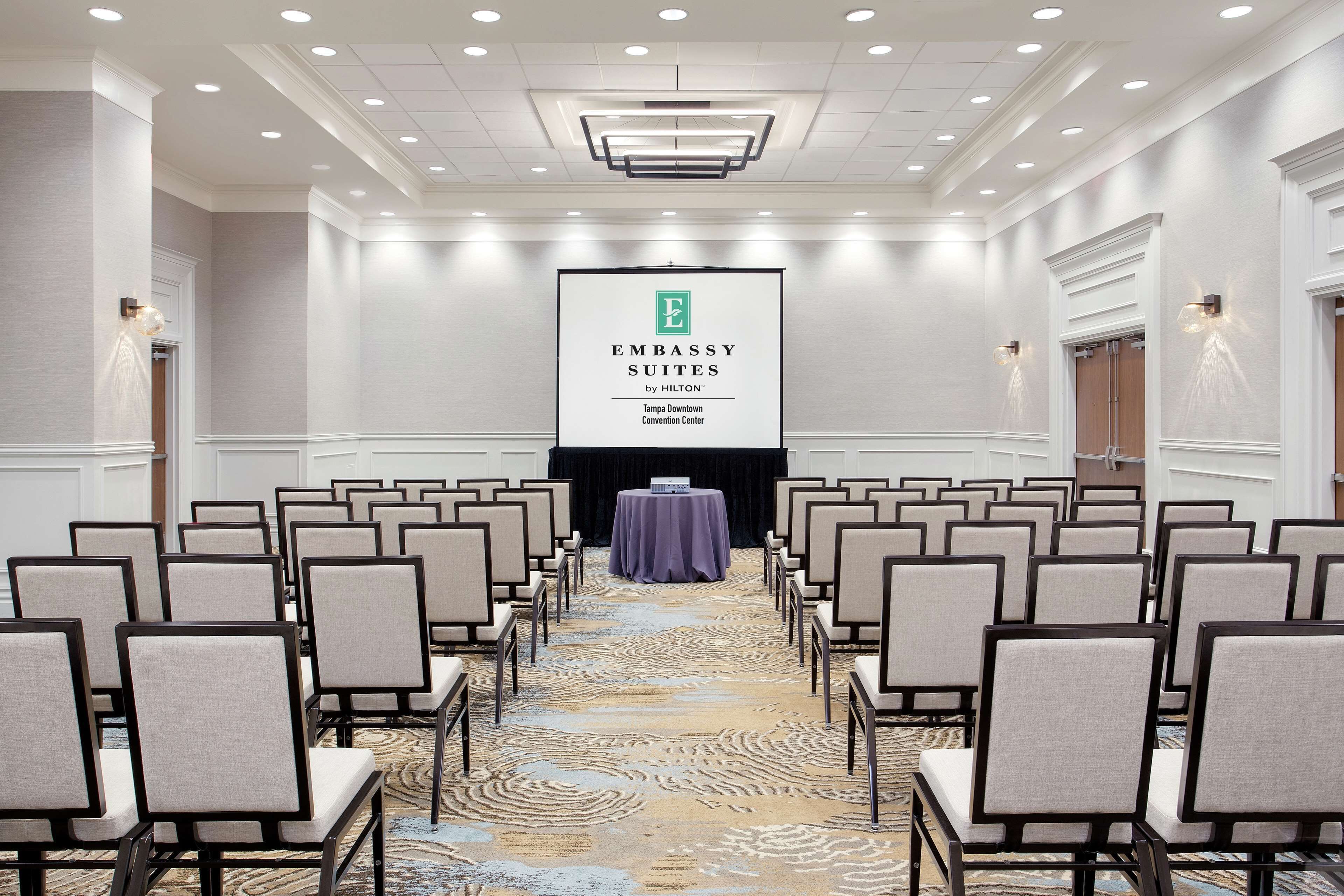 A conference room with rows of chairs, a stage with a presentation screen, and a carpeted floor.