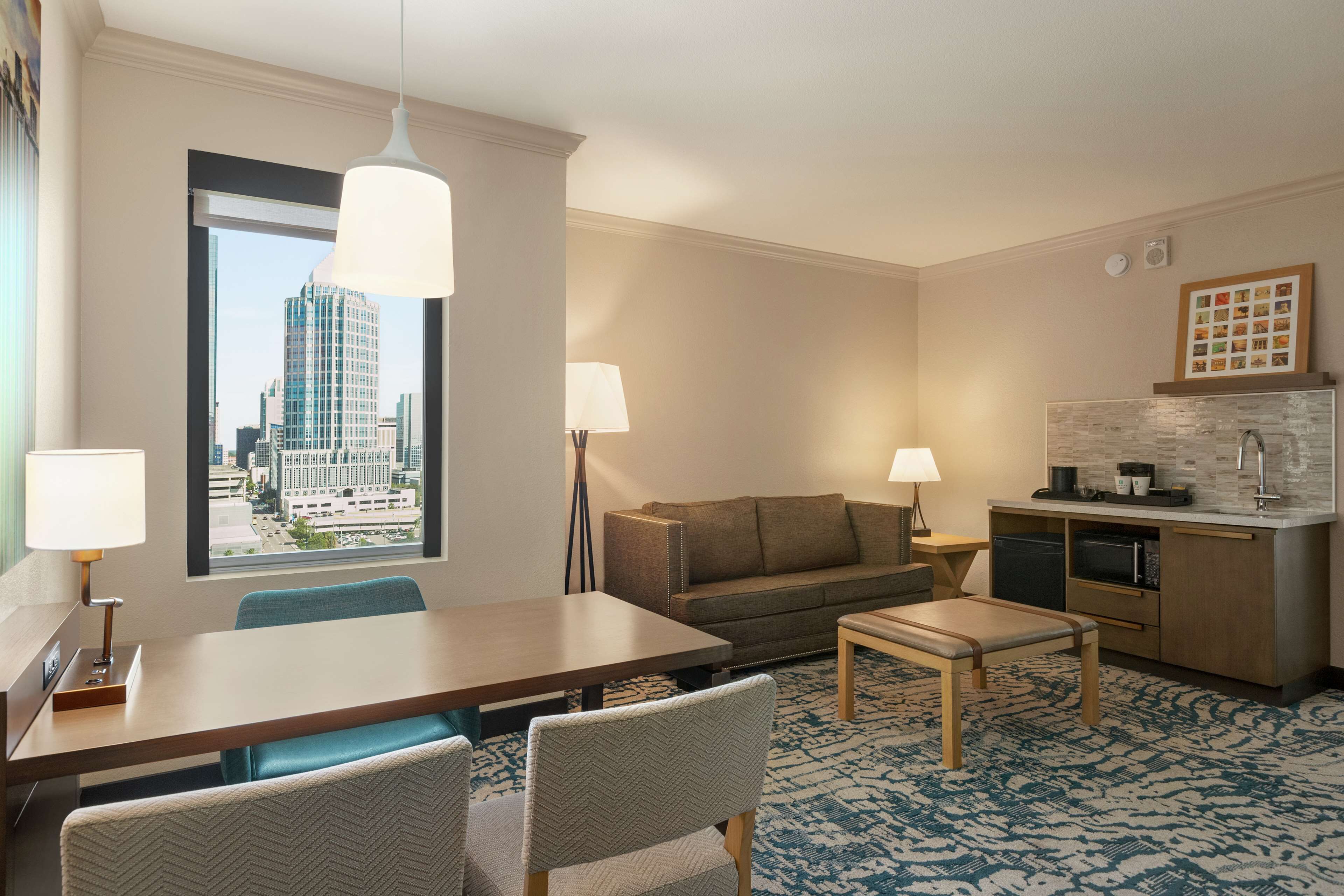 Ein Hotelzimmer mit Esstisch, zwei Stühlen, einer Couch, einem kleinen Tisch, einer Lampe, einer Küchenzeile und einem großen Fenster mit Blick auf die Stadt.