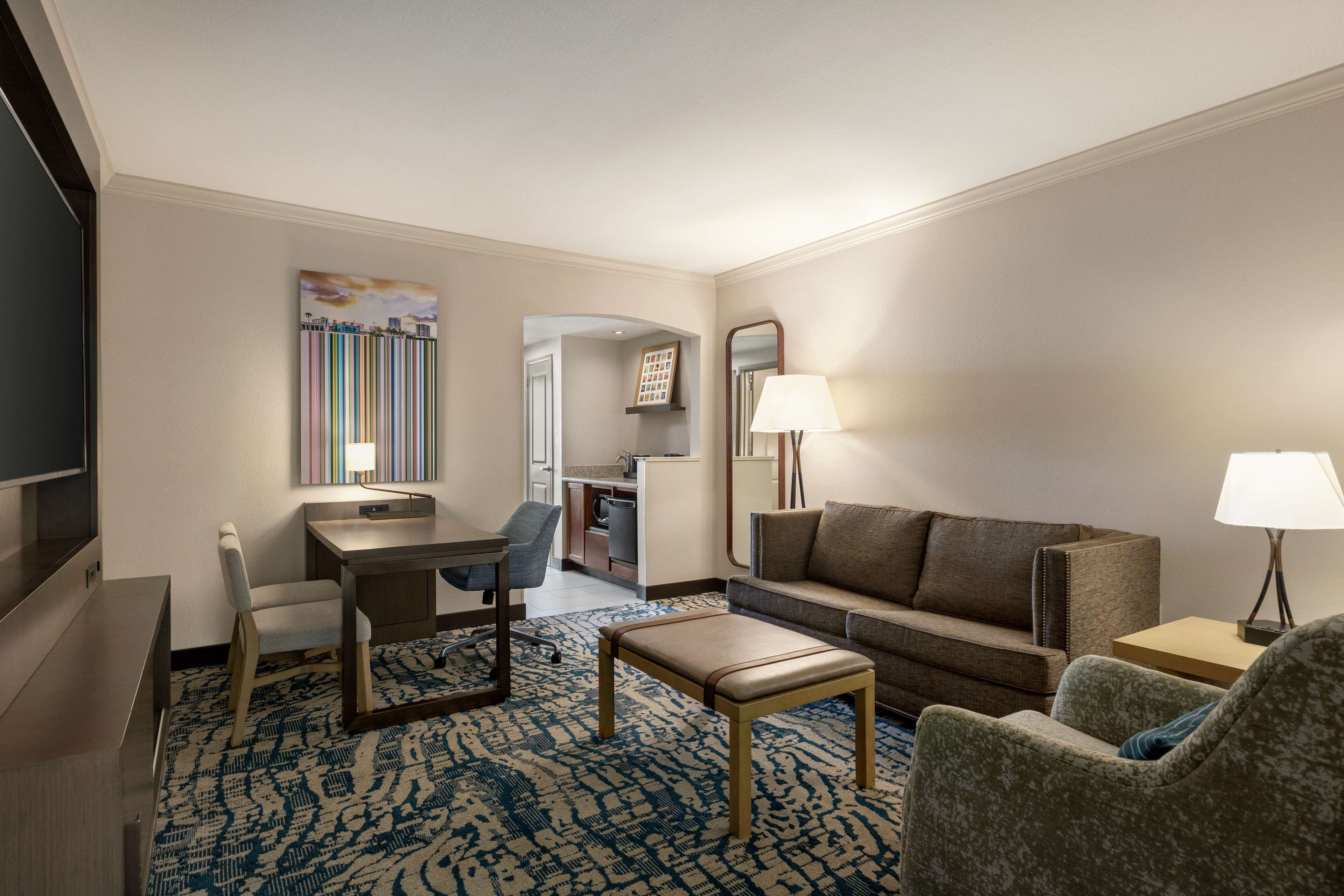 A hotel room with a flat-screen TV, a desk with a chair, a sofa, a patterned rug, and a dining area with a chair and table.